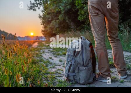 I piedi dell'uomo sono sulla pista nel campo contro il cielo con un tramonto in estate, circa i piedi dell'uomo è uno zaino. Il concetto di viaggio Foto Stock