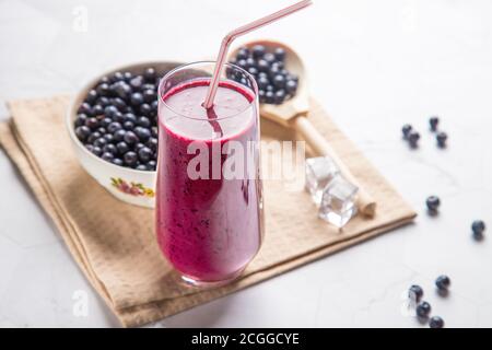 Dieta bere frullato di mirtillo in un bicchiere con tubo, piatto bianco con bacche su sfondo chiaro, con un tovagliolo beige, in un cucchiaio di legno-blu Foto Stock