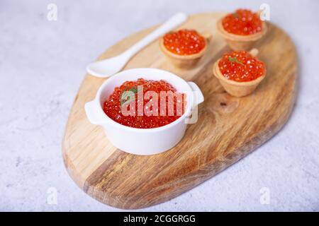 Caviale rosso (caviale di salmone) in una ciotola bianca su un asse di legno. Sullo sfondo sono i tartlet con caviale. Messa a fuoco selettiva, primo piano. Foto Stock