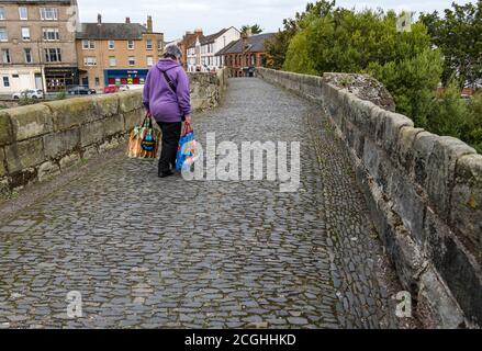 Donna che trasporta le borse della spesa della drogheria che cammina sopra il vecchio ponte romano acciottolato, Musselburgh, Lothian orientale, Scozia, Regno Unito Foto Stock