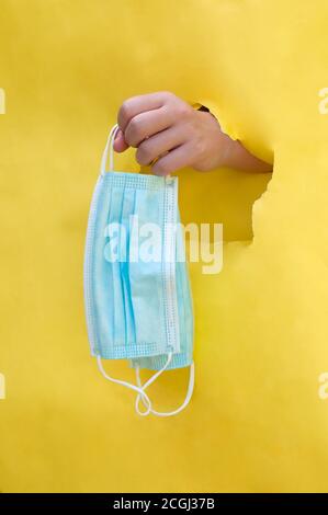 la foto verticale della mano attraverso il foro dentro lo sfondo giallo offre le maschere monouso blu Foto Stock