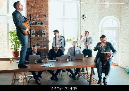 Cantare, dare concerti di musica, ballare. I colleghi sono felici e spensierati e si divertono in ufficio mentre i colleghi lavorano sodo e altamente concentrati. Concetto di divertimento, riposo, occupazione professionale. Foto Stock