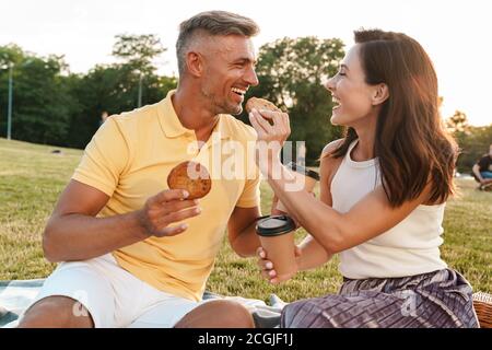 Ritratto di stupiti di mezza età giovane uomo e donna di bere il caffè da asporto e mangiare i cookie mentre è seduto sull'erba in posizione di parcheggio Foto Stock