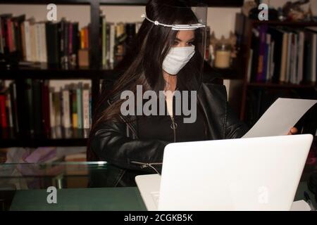 Donna che indossa maschera trasparente e copri bocca in ufficio accanto alla libreria e un computer portatile con vaporizzatore deodorante per ambienti legge un documento con concent Foto Stock