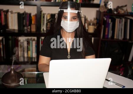 Donna di fronte a scrittura in avanti sulla macchina da scrivere in un ufficio con una grande libreria sullo sfondo mentre si indossa una maschera trasparente e protezione della bocca Foto Stock