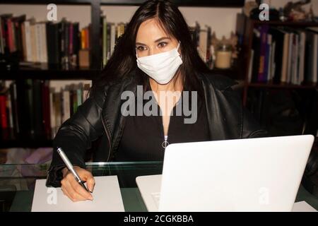 Donna dagli occhi ambra che indossa una copertura bianca della bocca che copre correttamente il naso e. bocca scrive su foglio bianco di lavoro di carta in un ufficio accanto al suo computer Foto Stock