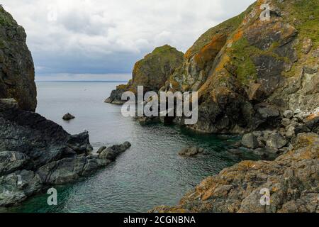 Immagine che mostra una stretta intrusione dell'oceano Atlantico tra i corpi di roccia costiera che formano una baia panoramica. Le rocce sono coperte di lichene di mare arancione e m. Foto Stock