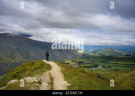 Un uomo indossa nero si erge su una collina per vedere una vista panoramica al punto di osservazione Arrow Junction durante l'estate ad Otago, Nuova Zelanda. Foto Stock