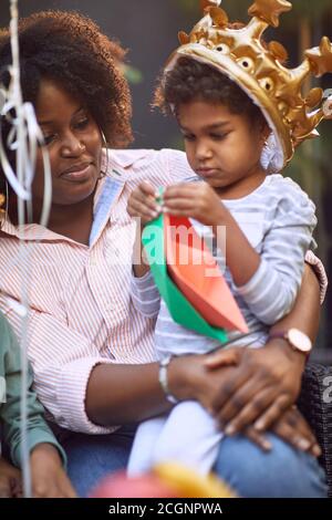 bambina afro-americana seduta nel grembo della sua mamma con corona gonfiabile gialla dorata sul suo odiato, giocando con barche di carta Foto Stock