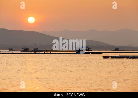 Una tranquilla comunità di case galleggianti su un lago al tramonto, gli agricoltori nutrendo pesce d'acqua dolce in gabbia di allevamento ittico, scenario rurale nella Thailandia occidentale. Foto Stock