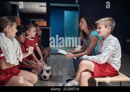 La squadra di calcio dei bambini e la loro allenatrice femminile in un armadietto stanza che parla di tattiche Foto Stock