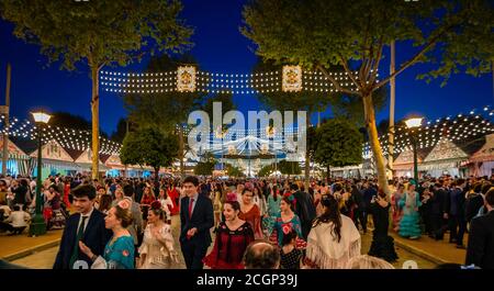 Spagnoli festeggiano in abiti tradizionali di fronte a marchee illuminati, casetas in serata, strada decorata, Feria de Abril, Siviglia, Andalusia Foto Stock