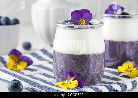 Sana colazione vegana di parfati di mirtillo fatta con frutti di bosco freschi, yogurt al cocco, budino di chia e fiori commestibili in bicchieri su tovagliolo su bianco Foto Stock