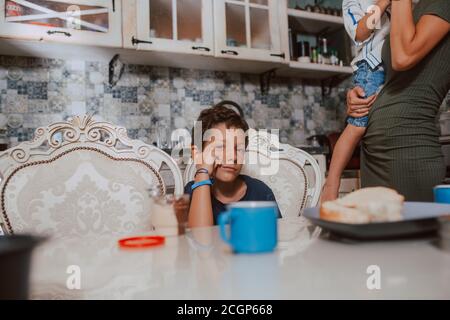 Una bella madre caucasica in un vestito con i suoi due figli è in piedi in cucina durante la prima colazione. Tempo di famiglia Foto Stock