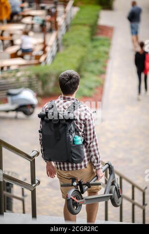 Giovane uomo caucasico in abiti casual con uno zaino porta uno scooter elettrico ripiegato in mano sulle scale di un edificio di ufficio. Uomo che porta Foto Stock