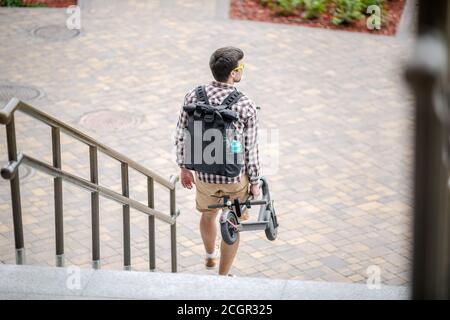 Giovane uomo caucasico in abiti casual con uno zaino porta uno scooter elettrico ripiegato in mano sulle scale di un edificio di ufficio. Uomo che porta Foto Stock