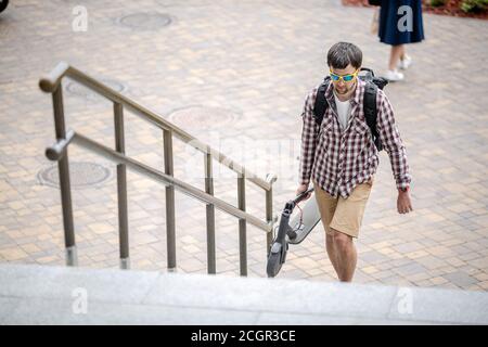 Giovane uomo caucasico in abiti casual con uno zaino porta uno scooter elettrico ripiegato in mano sulle scale di un edificio di ufficio. Uomo che porta Foto Stock