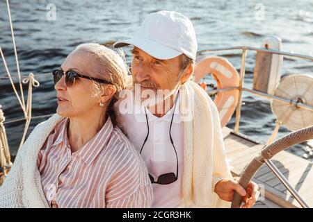 Coppia matura avvolta in plaid su yacht. Uomo anziano che tiene un volante e siede con sua moglie sullo yacht. Foto Stock