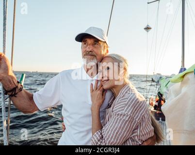 Affettuosa coppia senior abbracciando lo yacht e guardando in lontananza. Coppia ritirata in piedi insieme sulla barca a vela. Foto Stock
