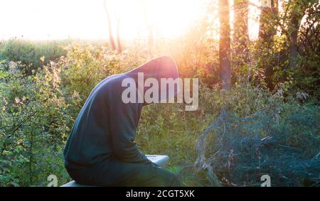 Un concetto di salute mentale di una moodia, figura agganciata, seduta su una panchina. Torna illuminato dal sole della sera. Foto Stock