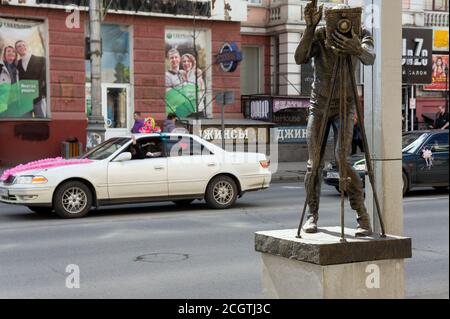 Monumento al fotografo sullo sfondo di auto da matrimonio su una strada della città. Sight si trova nel centro di Krasnoyarsk, Krasnoyarsk Terri Foto Stock