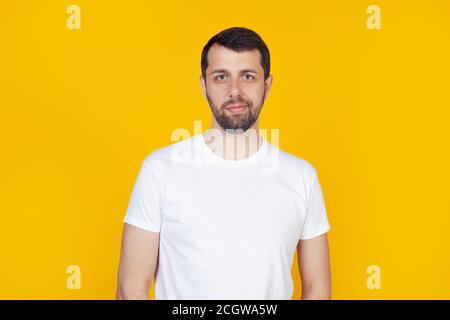 Giovane uomo con barba in una t-shirt bianca faccina sorridente guardando la macchina fotografica. Uomo positivo in piedi su sfondo giallo isolato. Foto Stock