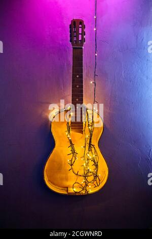 Vecchia chitarra in legno rotto decorata blu luminoso e luminoso ghirlando. Strumento musicale appeso alla parete di cemento. Decorazione di interni di Natale. FESTI Foto Stock
