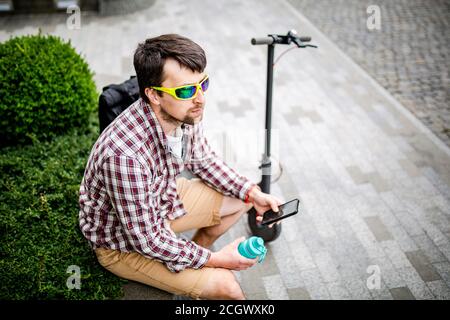 Moderno tema ecologico trasporto pulito. Un uomo che riposa su una panchina e tiene in mano uno smartphone dopo aver fatto un kick scooter eco-elettrico Foto Stock