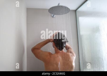 Uomo che prende una doccia che lava i capelli sotto l'acqua che cade dalla doccia a pioggia. Persona che doccia nello stile di vita domestico. Cura del corpo giovane adulto routine mattutina Foto Stock