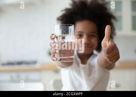 Ragazza biraciale che tiene un bicchiere d'acqua, mostrando i pollici verso l'alto gesto. Foto Stock