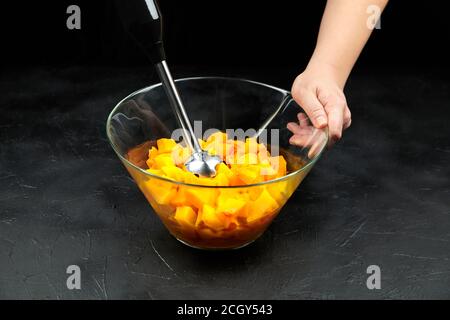 Pezzi di zucca e frullatore in un recipiente di vetro su un tavolo nero. Zucca tritata in piatto di insalata. Cucina di verdure, purea di zucca (purea) ricetta Foto Stock