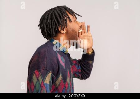 Vista laterale uomo africano con eleganti dreadlock gridando forte, annuncio di informazioni importanti, fobia. Studio al coperto isolato sulla parte posteriore grigia Foto Stock