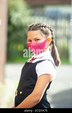 Una studentessa di dieci anni in una divisa scolastica con i capelli in trecce e indossando una maschera rosa. Foto Stock