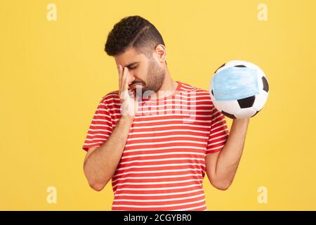 Profondamente sconvolto uomo bearded piangendo tenendo palla in mano maschera protettiva, vogliono visitare il campionato, partite chiuse a causa del coronavirus covid-19. IO Foto Stock