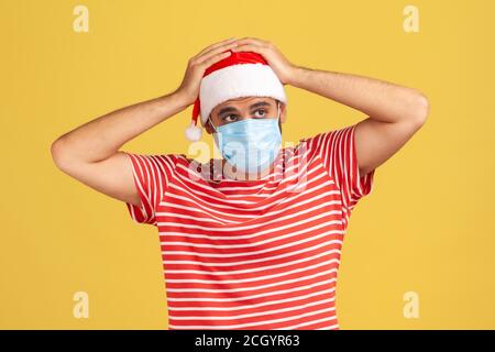 Infelice uomo triste in T-shirt rossa e cappello di babbo natale con maschera medica chirurgica sul viso che tocca la testa, emicrania, paura di contrarre il coronavirus. Interno Foto Stock