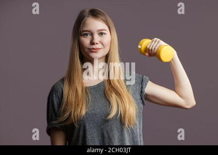 Attraente giovane donna fitness con capelli lisci biondi che indossa una t-shirt grigia che regge il manubri giallo. Corpo sano e forte. Foto Stock