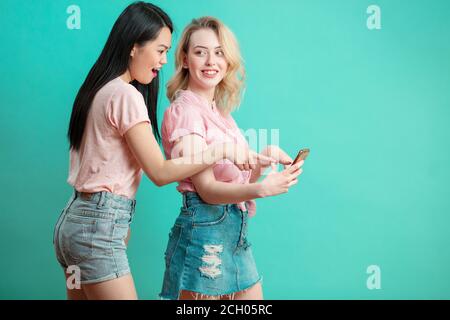 Due società multietnica bella giovane studente ragazze asiatici e caucasici in vestiti casual avendo divertimento prendendo selfie presso lo studio su sfondo blu Foto Stock