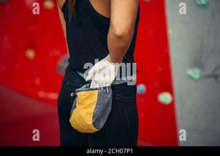 Vista posteriore di una donna irriconoscibile che applica una polvere di gesso di magnesio sulle mani da una borsa legata alla vita, da vicino sopra la parete di arrampicata rossa e bianca ba Foto Stock