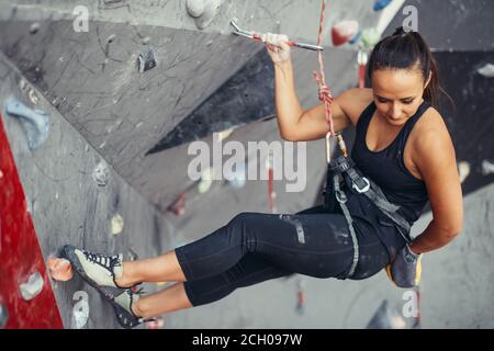 Montare sportivo ragazza studente appeso su ARIA utilizzando la fune di sicurezza e il sistema di cavi sulla roccia artificiale, guardando verso il basso, alla ricerca di un modo per ottenere il basso. Rosso e grigio a colori Foto Stock