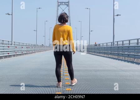 Vista posteriore di donna che cammina nel mezzo di una strada vuota verso il ponte Florianopolis. Ponte in acciaio sotto il bel cielo blu. Continuare a camminare conc Foto Stock