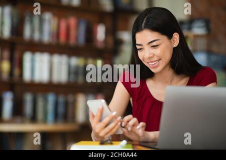 Bella donna asiatica che usa lo smartphone mentre lavora su un computer portatile In caffetteria Foto Stock