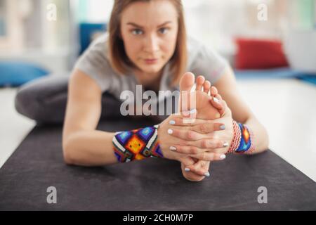 Vista frontale della giovane donna che guarda la macchina fotografica mentre si pratica yoga stretching esersice testa a ginocchio Forward Bend posa. Primo piano di mani e piedi Yogi. Foto Stock