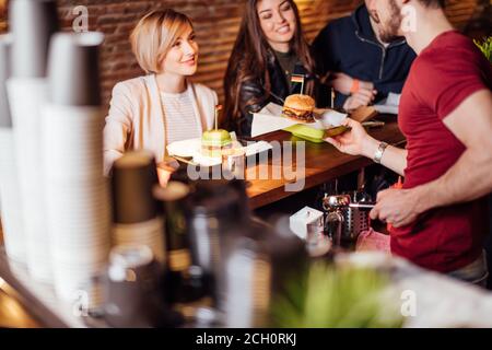 Gruppo di felici amici sorridenti che ordinano il cibo attraverso il barkeeper al banco in un ristorante moderno con stile loft, mattoni e tubi, interni Foto Stock