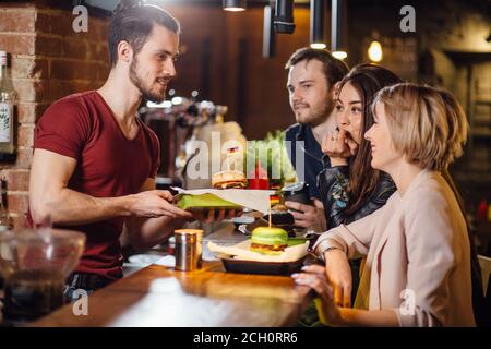 Gruppo di felici amici sorridenti che ordinano il cibo attraverso il barkeeper al banco in un ristorante moderno con stile loft, mattoni e tubi, interni, Foto Stock