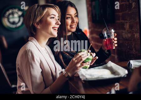 Due giovani ragazze caucasiche attraenti all'hamburger bar con pasto in mano, guardando la finestra curiosamente come se notassero intesting ragazzi fuori Foto Stock