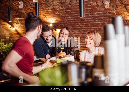 Gruppo di felici amici sorridenti che ordinano il cibo attraverso il barkeeper al banco in un ristorante moderno con stile loft, mattoni e tubi, interni Foto Stock