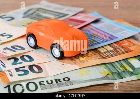 Auto giocattolo arancione su diverse banconote in euro. Concetto di acquisto, noleggio, assistenza, riparazione e costi assicurativi dell'automobile. Pianificazione delle spese. Vista frontale. Foto Stock