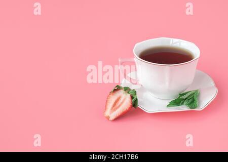 Tè con fragole e menta. Tazza di tè bianco su un piatto bianco con foglie di menta e fragole mature. Sfondo rosa. copyspace. Foto Stock