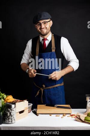 Studio di caucasico felice uomo d'affari in grembiule blu abiti da lavoro che decidono di dare da mangiare ai suoi ospiti e affilare il coltello in cucina contro Foto Stock