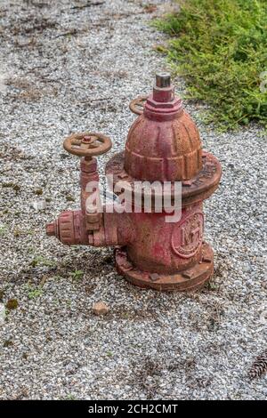 Vecchio idrante rosso fuoco parzialmente nel terreno con molti strati di peeling vernice rustica e indossata ancora in servizio con due valvole a saracinesca per la pressione dell'acqua Foto Stock